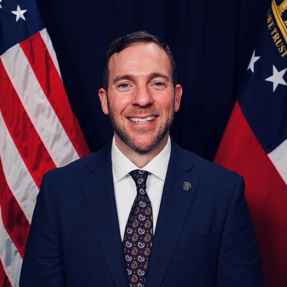 Man in Navy Blue suit smiles in front of Georgia and American flags